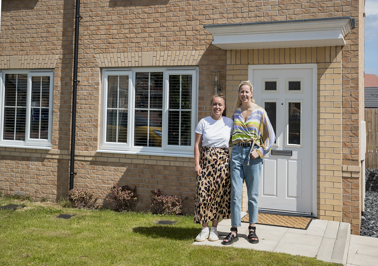 A front-view shot of a couple standing outside their new home, they have wide smiles on their faces.